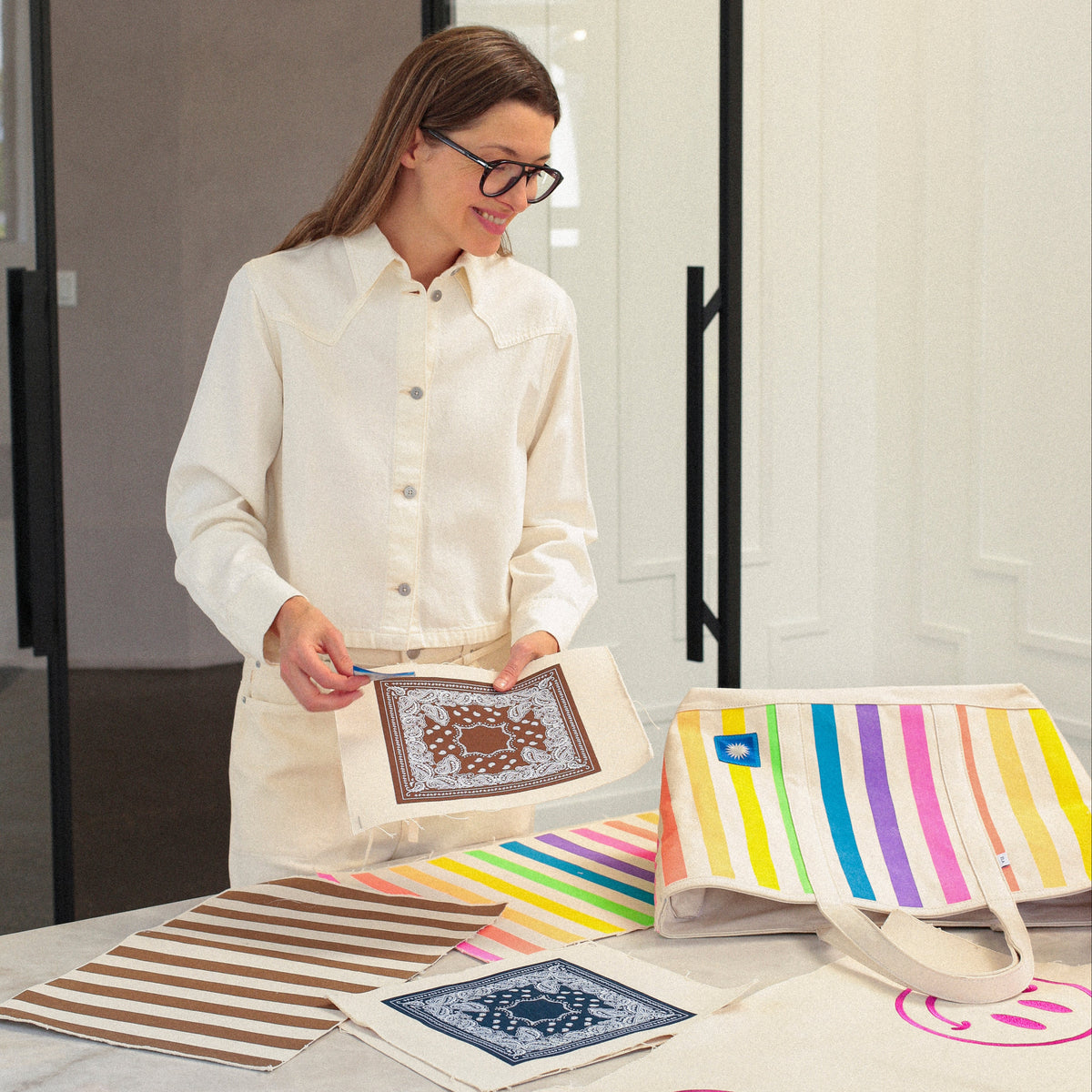 Founder of ELA, in a cream shirt holds a printed canvas tote bag with a geometric pattern, standing beside a table displaying multiple colorful striped and printed canvas tote bags in a bright, modern room.